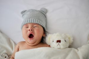 Bébé avec un bonnet gris avec oreilles en train de bailler dans son lit avec son nounours blanc