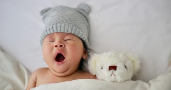 Bébé avec un bonnet gris avec oreilles en train de bailler dans son lit avec son nounours blanc
